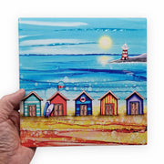 5 Beach Huts and Lighthouse ceramic trivet held in hand showing full design with five colourful beach huts and red and white striped lighthouse