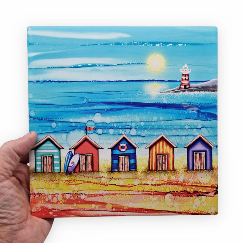5 Beach Huts and Lighthouse ceramic trivet held in hand showing full design with five colourful beach huts and red and white striped lighthouse