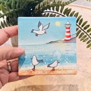 Seagull and Lighthouse ceramic coaster held in hand showing full coastal scene with seagulls, beach and red and white striped lighthouse
