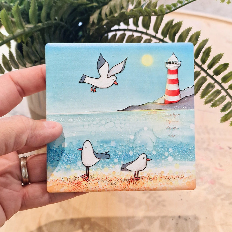 Seagull and Lighthouse ceramic coaster held in hand showing full coastal scene with seagulls, beach and red and white striped lighthouse