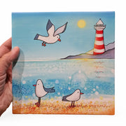 Seagull and Lighthouse ceramic trivet held in hand showing full coastal design with sandy beach and sparkling sea