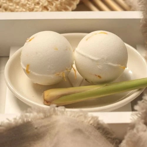 Two white Clovelly Cedarwood and Lemongrass aromatherapy bath bombs on a cream ceramic dish with a fresh lemongrass stalk, displayed on a white wooden bath tray