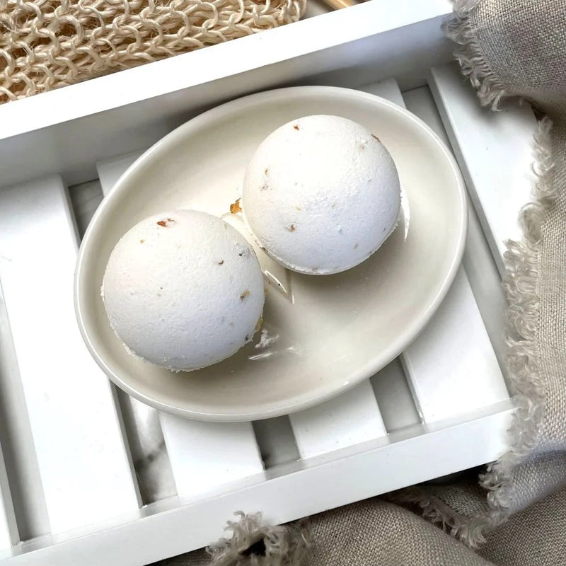 Two white Clovelly aromatherapy bath bombs with jasmine flowers on a cream ceramic dish, displayed on a white wooden bath tray