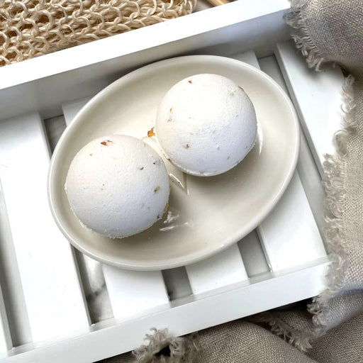 Two white Clovelly aromatherapy bath bombs with jasmine flowers on a cream ceramic dish, displayed on a white wooden bath tray