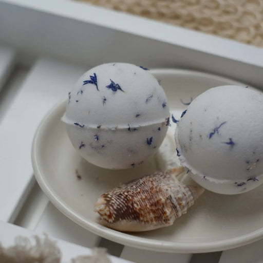 Two white Clovelly Ocean Breeze aromatherapy bath bombs with blue cornflower petals on a cream ceramic dish with a seashell, displayed on a white wooden bath tray