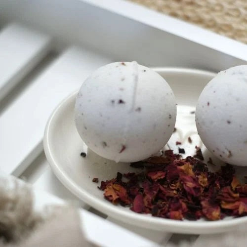 Two white Clovelly Rose Geranium aromatherapy bath bombs on a cream ceramic dish with dried rose petals, displayed on a white wooden bath tray