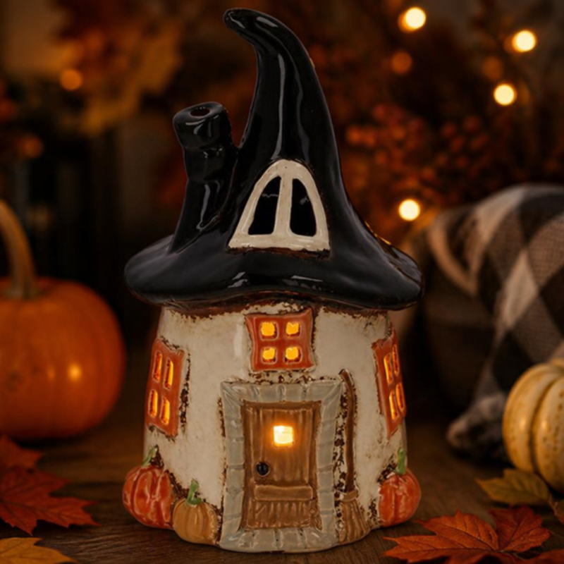 Village Pottery Witch Round House Tealight Holder with glowing windows, witch’s hat roof, chimney, broom and pumpkins among autumn leaves.