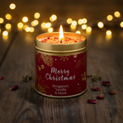 Merry Christmas candle tin lit with a warm glowing flame, styled on a rustic wooden surface with golden bokeh fairy lights in a cosy festive setting