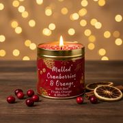 Mulled Cranberries & Orange Christmas candle tin lit with a warm glowing flame, styled on a rustic wooden surface with golden bokeh fairy lights and cranberries and orange slices in a cosy festive setting