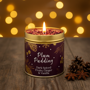 Plum Pudding Christmas candle tin lit with a warm glowing flame, styled on a rustic wooden surface with golden bokeh fairy lights in a cosy festive setting
