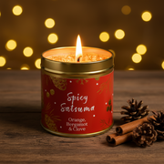 Spicy Satsuma Christmas candle tin lit with a warm glowing flame, styled on a rustic wooden surface with golden bokeh fairy lights and pine cones in a cosy festive setting