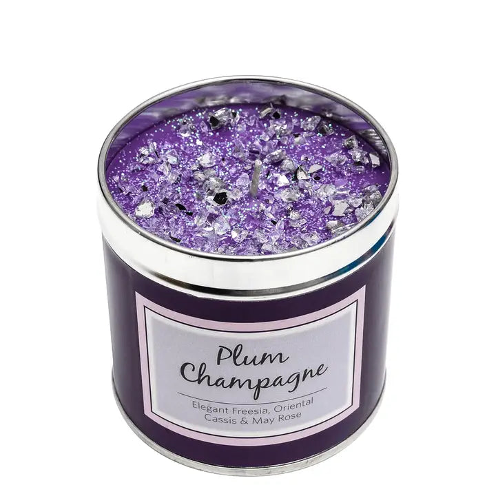 Plum Champagne scented candle in a tin with purple sparkly topping, label reading "Plum Champagne — Elegant Freesia, Oriental Cassis & May Rose", white background