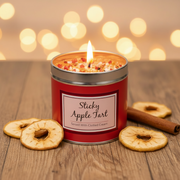 Sticky Apple Tart scented candle lit and glowing in a tin with sparkly topping, styled on a wooden surface with dried apple slices and cinnamon stick, warm bokeh background