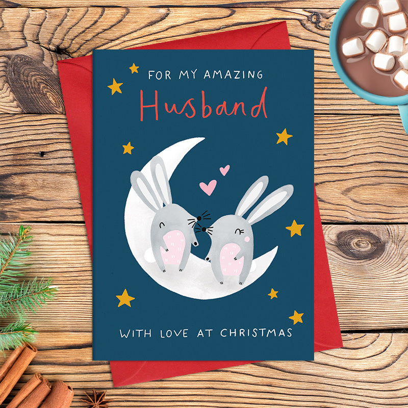 Amazing Husband Christmas card featuring two illustrated rabbits sitting together on a crescent moon, surrounded by stars and hearts, with the message ‘With love at Christmas’