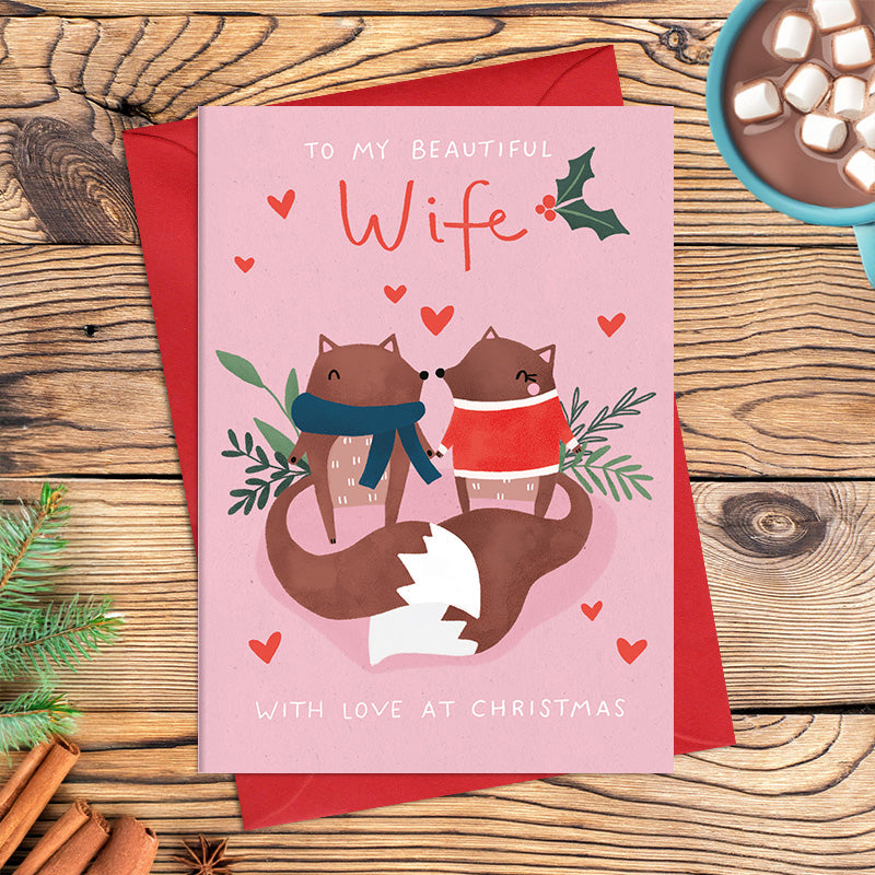 Beautiful Wife Christmas card featuring two illustrated foxes sitting nose to nose in cosy winter clothing, surrounded by hearts and foliage, with the message ‘With love at Christmas’.