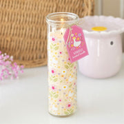 Lit Daisy and Duck vanilla scented glass tube candle with pink gift tag on a white surface with wicker basket and pink flowers in background