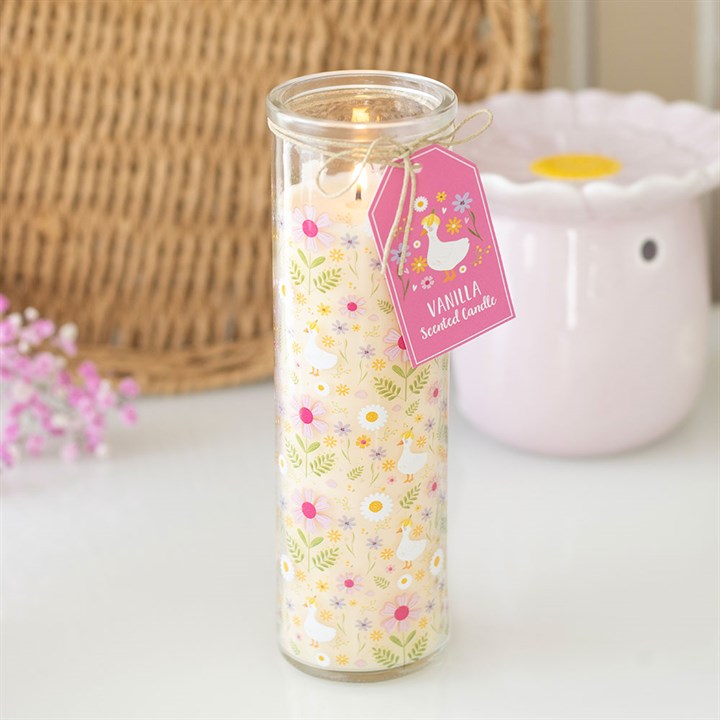Lit Daisy and Duck vanilla scented glass tube candle with pink gift tag on a white surface with wicker basket and pink flowers in background