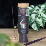 Dark Forest Wild Berry Incense Sticks, lifestyle shot of glass tube with mushroom and crescent moon label standing upright on wooden surface with incense stick burning in wooden moon phase holder, plant in background