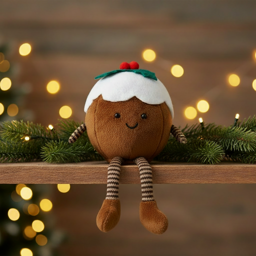 Plush Christmas pudding soft toy with smiley face, stripy legs and holly sprig, styled in a warm cosy festive scene with soft golden fairy lights