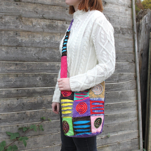 Patchwork spiral appliqu\u00e9 cross body bag worn across the body by a model in a cream cable knit jumper, standing against a wooden fence outdoors