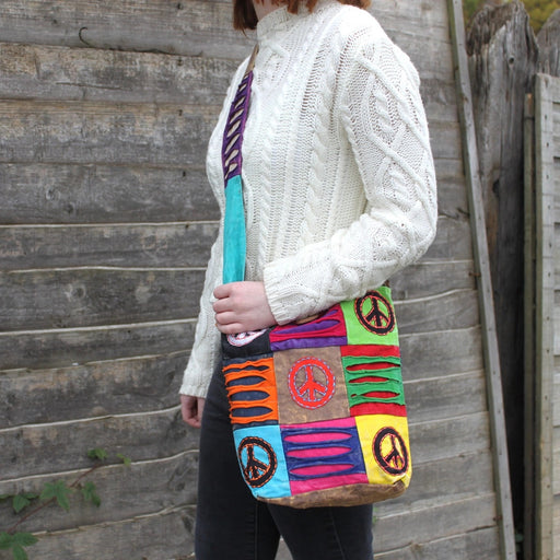 Patchwork peace sign hobo cross body bag worn across the body by a model in a cream cable knit jumper, standing against a wooden fence outdoors