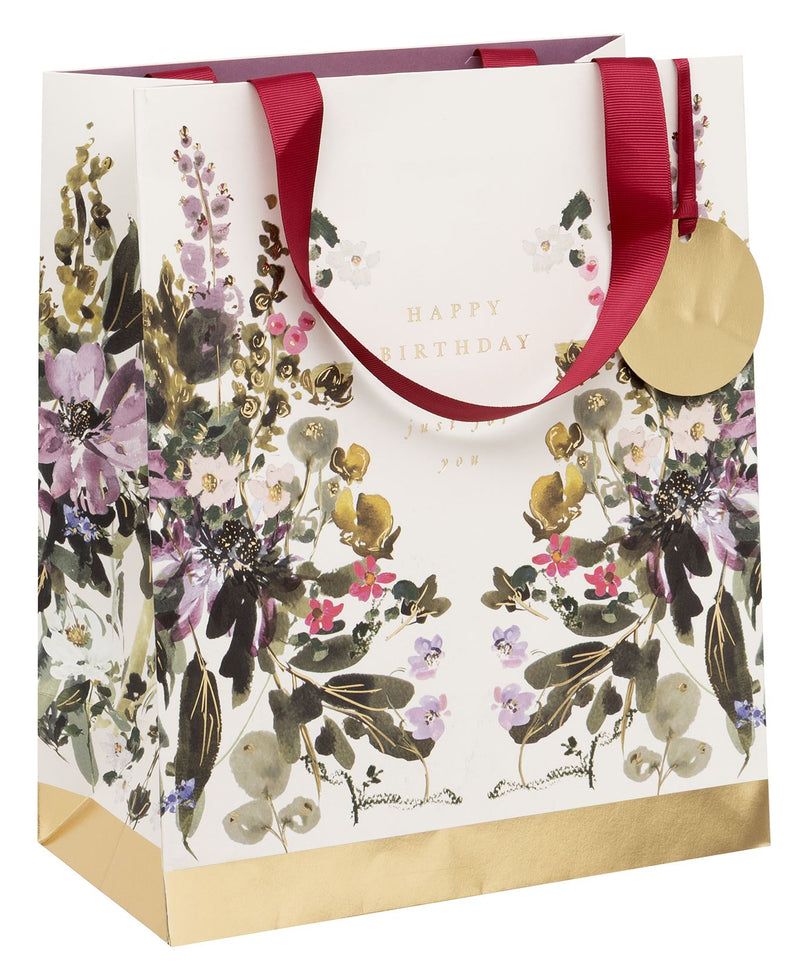 Stephanie Dyment Sunlit Harvest Large Happy Birthday Gift Bag with watercolour botanical print in purples, pinks and greens, gold foil accents, "Happy Birthday — just for you" in gold foil lettering, burgundy ribbon handles, gold foil base and circular gold gift tag on white background
