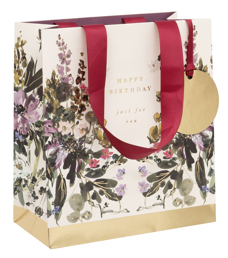 Stephanie Dyment Sunlit Harvest Medium Happy Birthday Gift Bag with watercolour botanical print in purples, pinks and greens, gold foil accents, "Happy Birthday — just for you" in gold foil lettering, burgundy ribbon handles, gold foil base and circular gold gift tag on white background