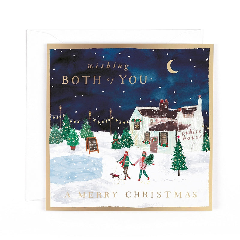Both of You Christmas card featuring a couple walking hand in hand with a dog beside a snowy village pub, Christmas trees and strings of lights beneath a crescent moon.