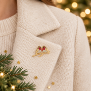 Gold plated crystal two robins brooch pinned on a cream knitted jacket lapel, surrounded by gold star confetti and evergreen sprigs with warm golden bokeh fairy lights in the background
