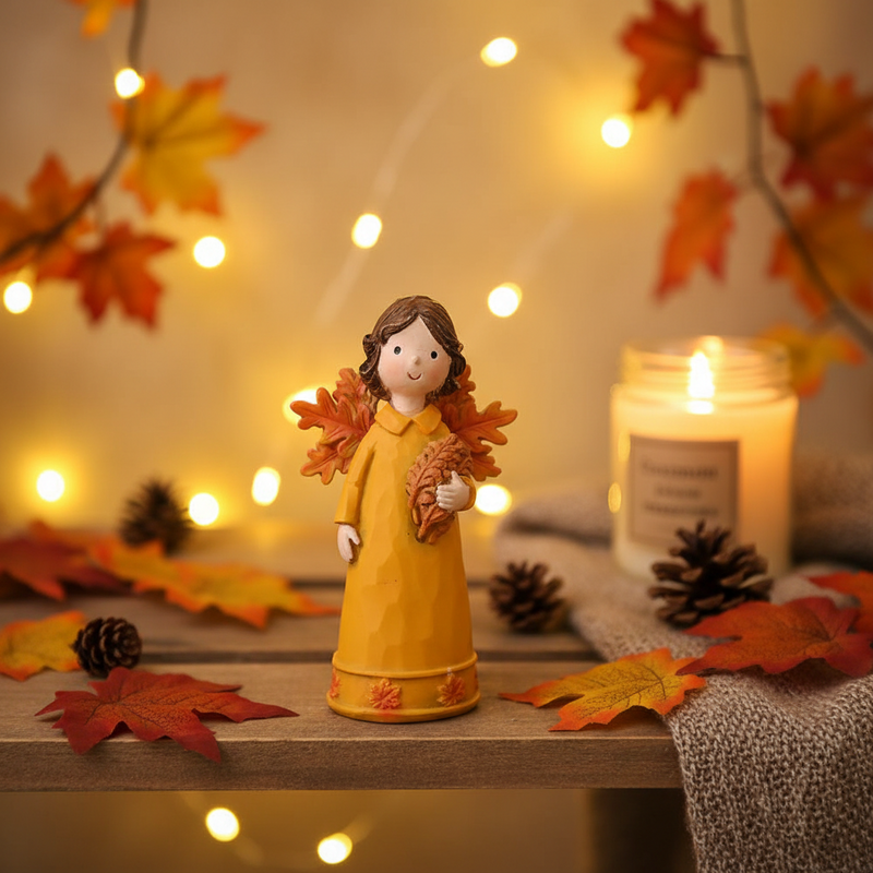 A small autumn fairy figurine in a mustard yellow dress with orange autumn leaf wings, displayed on a wooden surface in a warm cosy autumnal indoor setting with golden amber tones and soft bokeh fairy lights in the background