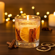 A clear gel candle in a glass tumbler with orange slices and cinnamon sticks suspended inside and a single wick at the top, displayed on a wooden surface in a warm cosy festive setting with rich dark tones and soft golden bokeh candlelight in the background