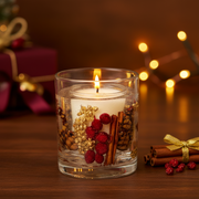 A clear gel candle in a glass tumbler with red berries, gold berries, cinnamon sticks and pinecones suspended inside and a single wick at the top, displayed in a warm cosy Christmas setting with deep burgundy and gold tones and soft golden bokeh fairy lights in the background