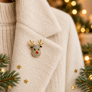 Gold plated crystal reindeer brooch pinned on a cream knitted jacket lapel, surrounded by gold star confetti and evergreen sprigs with warm golden bokeh fairy lights in the background