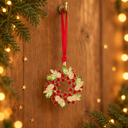 Gold plated crystal wreath decoration hanging by a long red ribbon threaded through a rustic gold hook, surrounded by evergreen sprigs and gold star confetti with warm golden bokeh fairy lights in the background