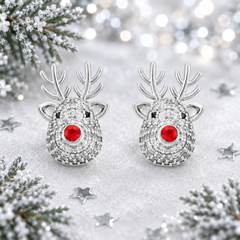 Pair of rhodium plated crystal reindeer earrings laid on a soft silvery white surface, surrounded by silver star confetti and frosted evergreen sprigs with cool bokeh fairy lights in the background
