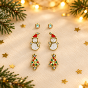 Trio of gold plated crystal, snowman and tree stud earrings laid face-up on a soft cream surface, surrounded by gold star confetti and evergreen sprigs with warm golden bokeh fairy lights in the background