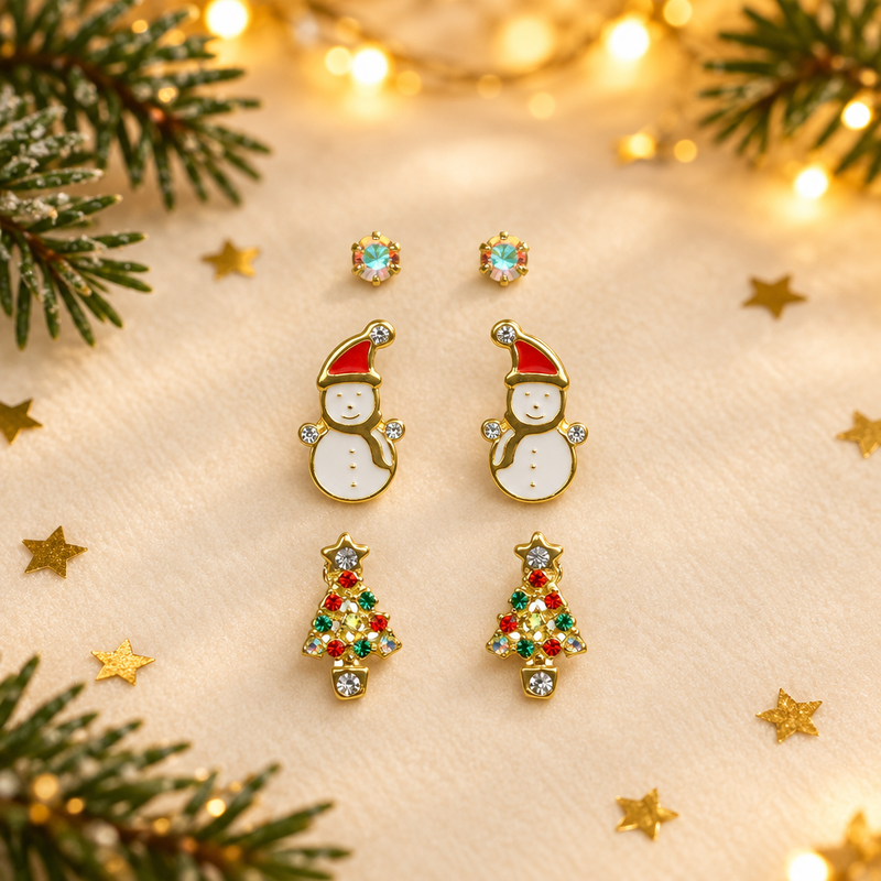 Trio of gold plated crystal, snowman and tree stud earrings laid face-up on a soft cream surface, surrounded by gold star confetti and evergreen sprigs with warm golden bokeh fairy lights in the background