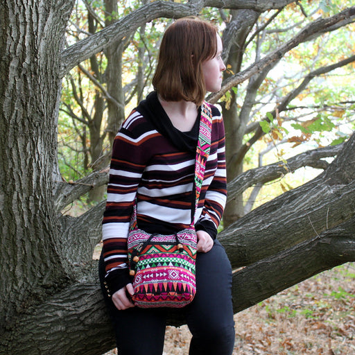 Pink Aztec print jacquard sling bag worn across the body by a model leaning against a tree in a woodland setting