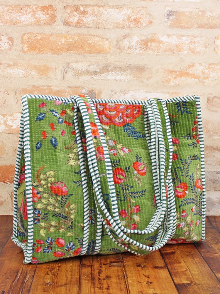 Quilted velvet tote bag in vibrant floral print with striped handles, sitting on a wooden surface against a stone wall