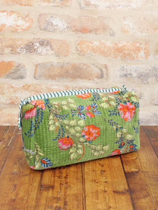 Quilted velvet wash bag in a vibrant floral print with striped zip trim, sitting on a wooden surface against a stone wall