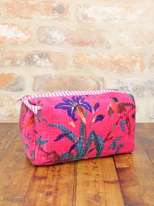 Quilted velvet wash bag in bold tropical floral print with striped zip trim, sitting on a wooden surface against a stone wall