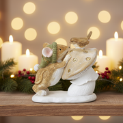 A decorative figurine of a mouse wearing a green hat leaning against a toadstool with a small bird perched on top, displayed on a shelf against a warm cosy Christmas background with soft bokeh lights