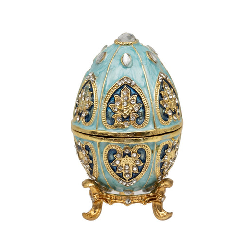 Turquesa Egg Trinket Box closed, turquoise egg-shaped trinket box with gold-tone detailing, heart-shaped dark blue panels with floral motifs and crystal-effect stones, large crystal-effect stone at top, on decorative gold-tone footed stand, on white background