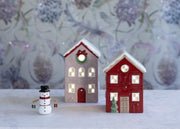 Conifer LED Cottage and Juniper LED Cottage ceramic LED houses glowing with warm light displayed alongside a snowman ornament on a white surface with floral background