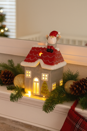 Santa on the Rooftop LED House glowing ceramic LED house with Santa on the rooftop displayed on a mantelpiece with pine garland pine cones and gold baubles