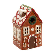 Gingerbread House Tealight Holder, warm brown ceramic gingerbread house with white icing-style scalloped snow-dusted roof tiles and red trim, green wreath gable window, white-framed grid windows with icing borders, dark arched door with candy cane arch, snowflake motifs and white scroll flourishes, on white background