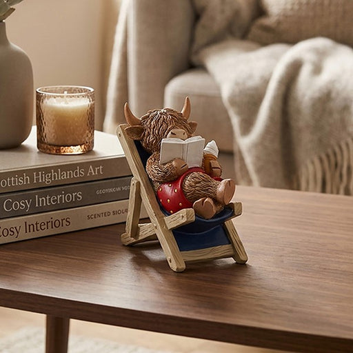 Highland cow figurine sitting in a blue deckchair reading a book and holding an ice cream, dressed in swimming trunks with shaggy fur and curved horns