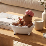 Capybara relaxing in a bathtub, wearing a shower cap and holding a cocktail, displayed on a wooden table.