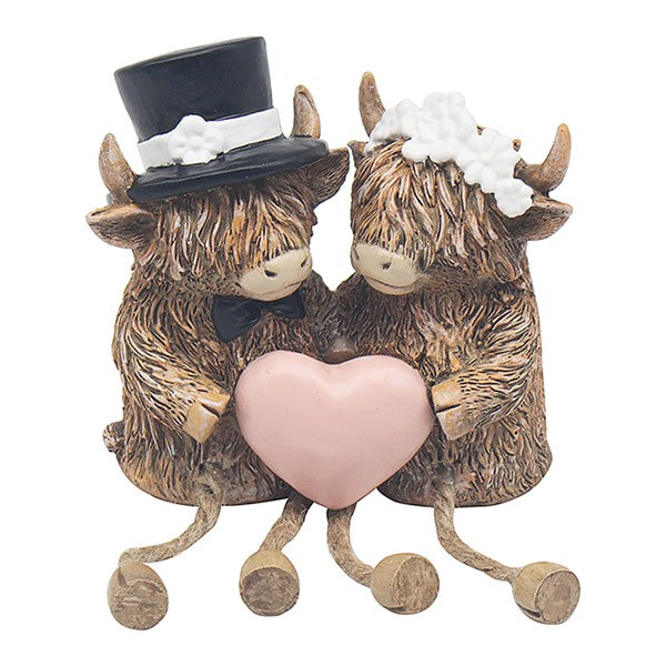 Highland cow bride and groom dangley legs figurine, groom in black top hat and bow tie and bride in white floral headpiece, holding a pink heart, on a white background