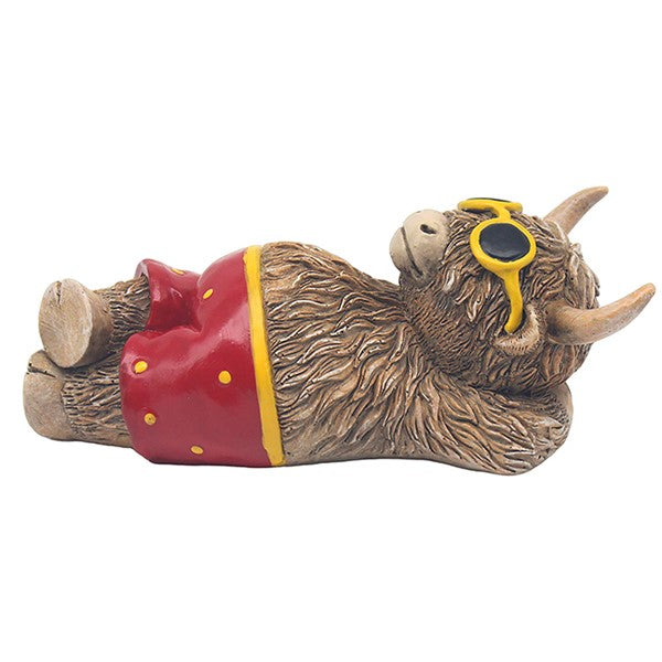 Happy Highland Cow Beach Time figurine – shaggy highland cow lying down wearing red polka dot swimming trunks and yellow sunglasses, with curved horns